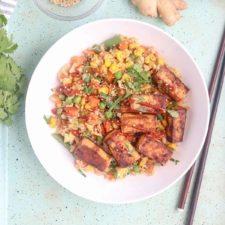 A white bowl filled with tofu cauliflower rice, colorful vegetables, and golden-brown tofu slices, garnished with chopped herbs. Chopsticks rest beside the bowl on a light blue speckled surface.