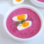 A bowl of bright pink Cold Beet Soup is garnished with a sprig of dill and two halves of a soft-boiled egg. The vibrant soup is served in a white bowl on a light surface.