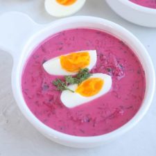 A bowl of bright pink Cold Beet Soup is garnished with a sprig of dill and two halves of a soft-boiled egg. The vibrant soup is served in a white bowl on a light surface.