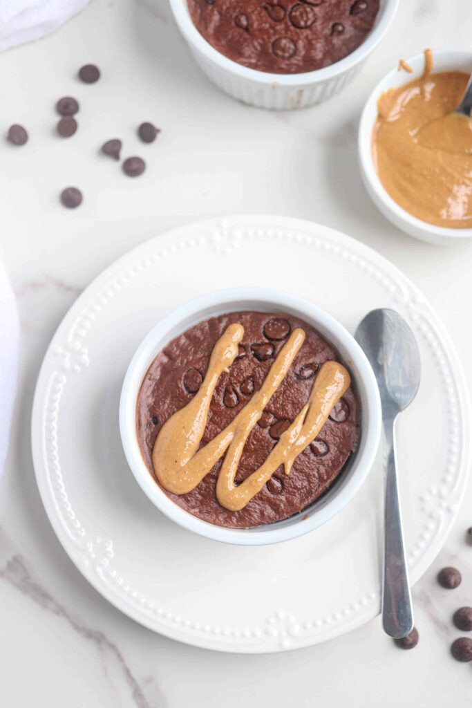 chocolate baked oats in a white ramekin on a white plate with chocolate chips and drizzled with peanut butter.