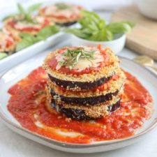 A stack of crispy Air Fryer Eggplant Parm slices layered with tomato sauce and melted cheese, garnished with fresh basil, sits on a plate with extra tomato sauce.
