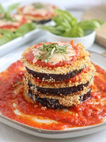 A stack of crispy Air Fryer Eggplant Parm slices layered with tomato sauce and melted cheese, garnished with fresh basil, sits on a plate with extra tomato sauce.