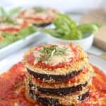 A plate with a stack of breaded, air-fried eggplant slices topped with tomato sauce, melted cheese, and fresh basil. Text above reads Air Fryer Eggplant Parm. In the background, more eggplant and basil are visible.