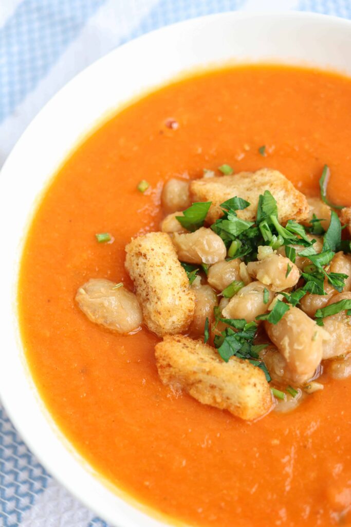 a white bowl of creamy tomato bean soup with croutons and chopped herbs on top on a blue and white striped kitchen towel, closeup