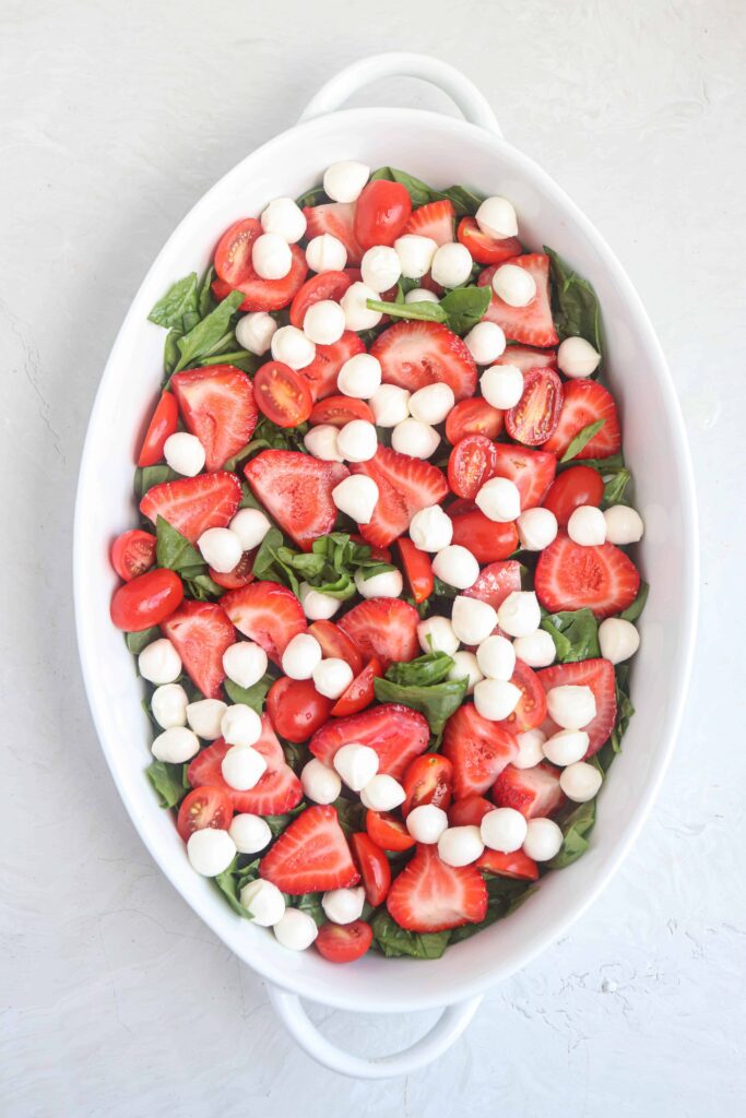 an oval dish with chopped baby spinach, strawberries and cherry tomatoes and mozzarella balls