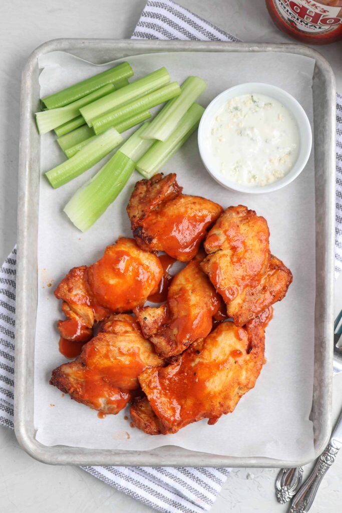 air fryer buffalo chicken thighs on a baking tray served on white paper with a side of celery sticks and blue cheese dressing