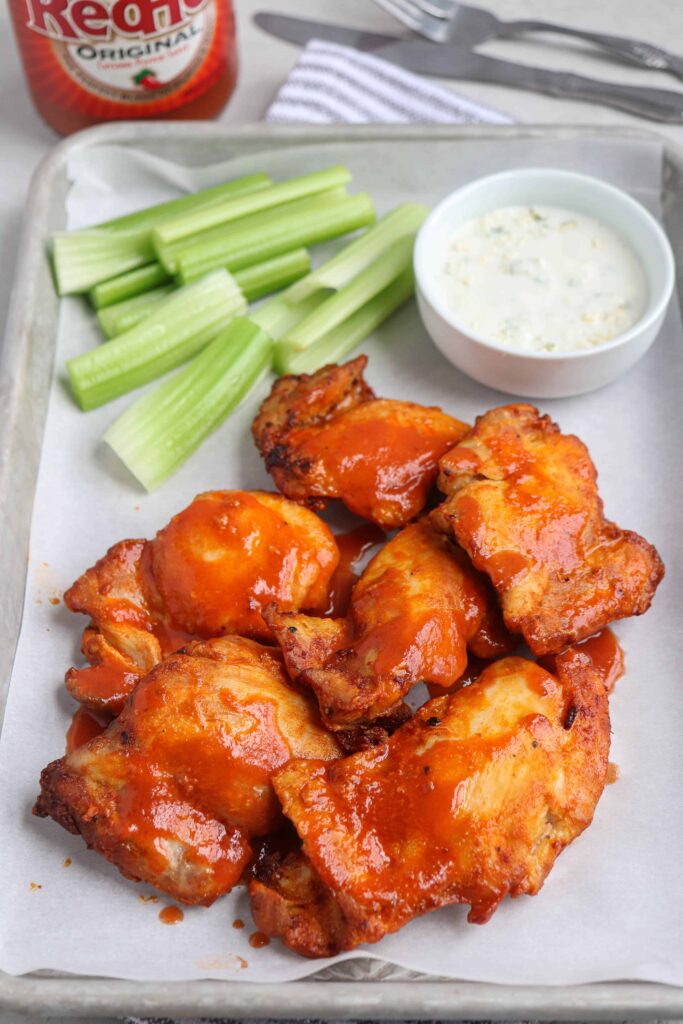 air fryer buffalo chicken thighs on a baking tray served on white paper with a side of celery sticks and blue cheese dressing.