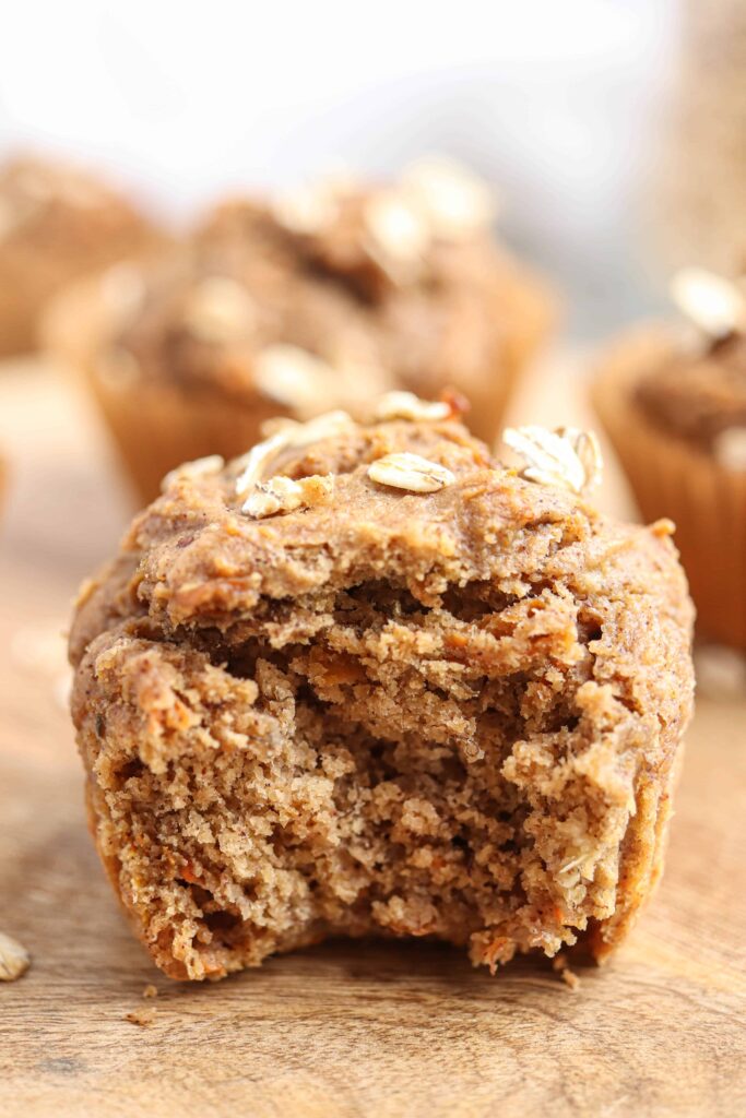 bite shot of banana carrot oat muffins on a round wooden board on light gray surface.