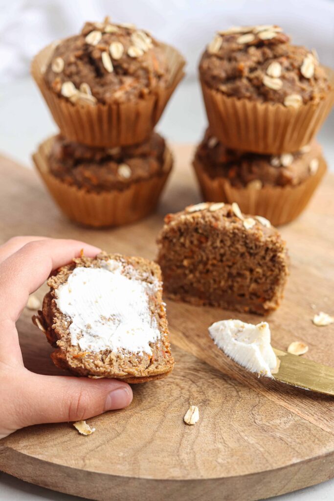 banana carrot oat muffins on a round wooden board on light gray surface, one muffin being buttered.