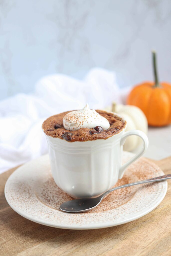 pumpkin mug cake with a dollop of whipped cream and cinnamon in a white teacup on a saucer