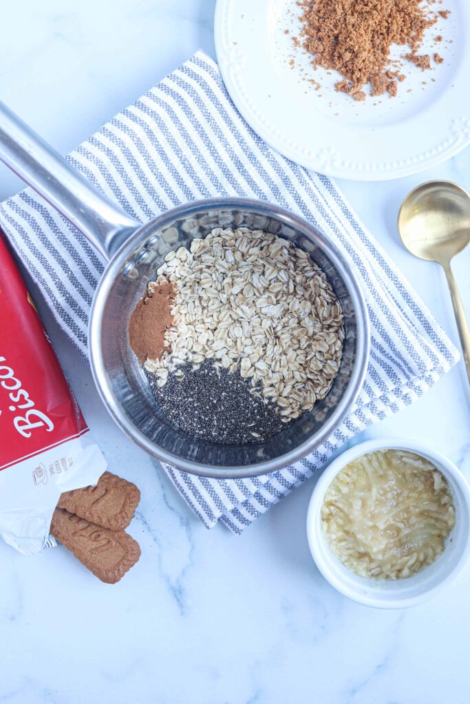oats, chia and cinnamon in a stainless steel pot on a striped white and gray kitche towel surrounded by Biscoff cookies, a mashed banana in a white ramekin and a golden spoon