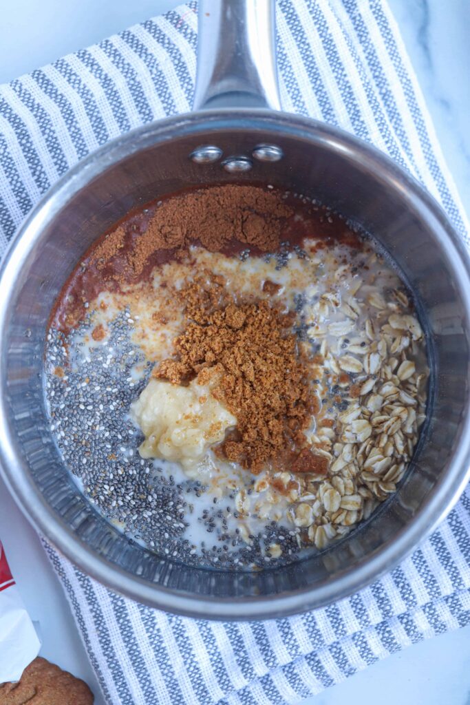 oats, chia seends, mashed banana, cinnamon and crushed Biscoff cookie in a stainless steel saucepan on a striped white and gray kitchen towel