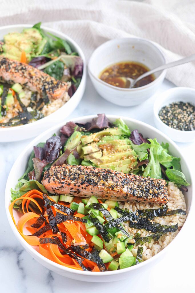 a white bowl with furikake salmon on the bed of greens, avocado, carrots, brown rice and nori on white marble surface