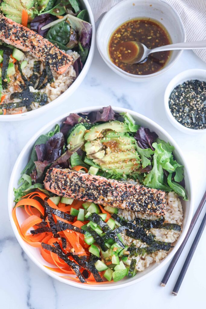a white bowl with furikake salmon on the bed of greens, avocado, carrots, brown rice and nori on white marble surface
