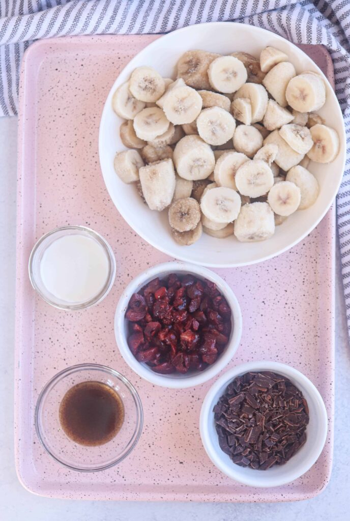 frozen bananas, cherries, chocolate pieces, vanilla and milk in bowls on a pink tray.