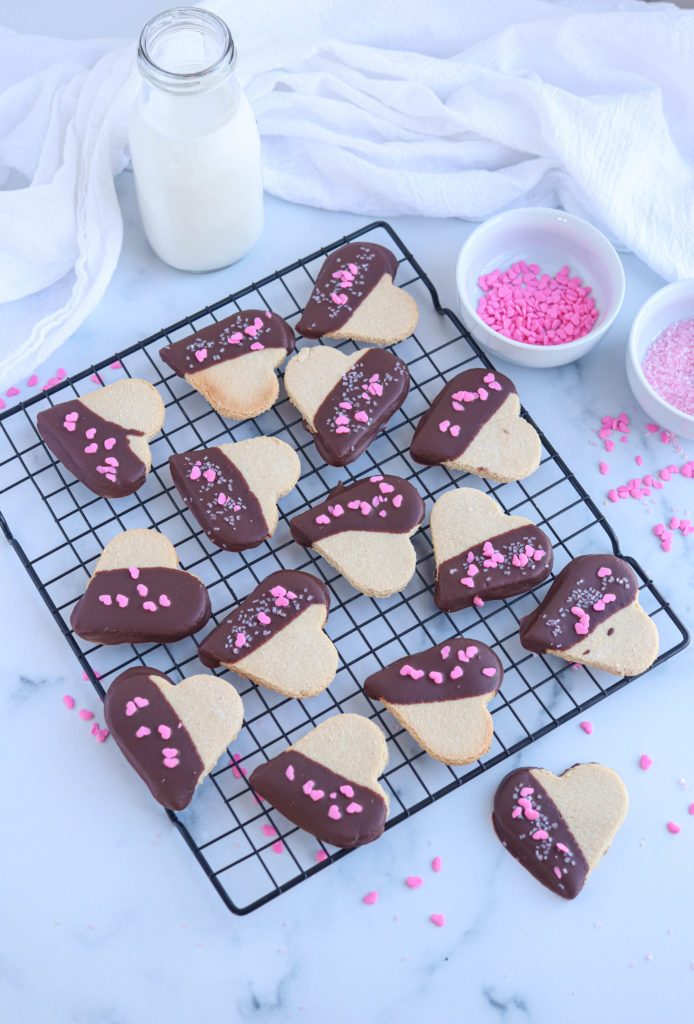 heart shaped chocolate dipped vegan shortbread cookies with pink heart sprinkles on a black cooling rack