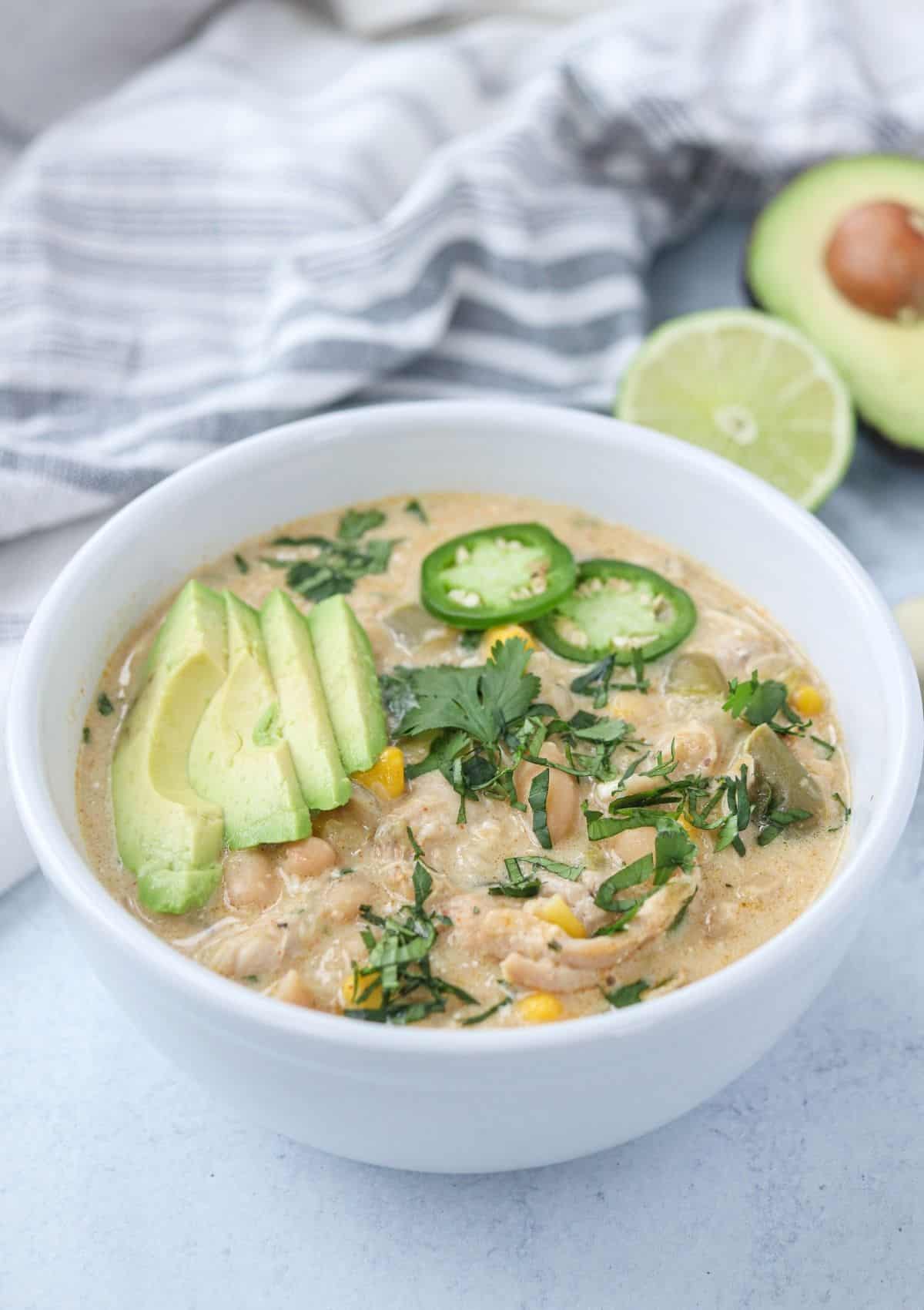 green chicken chili in a white bowl topped with avocado slices, jalapeno peppers and chopped cilantro.