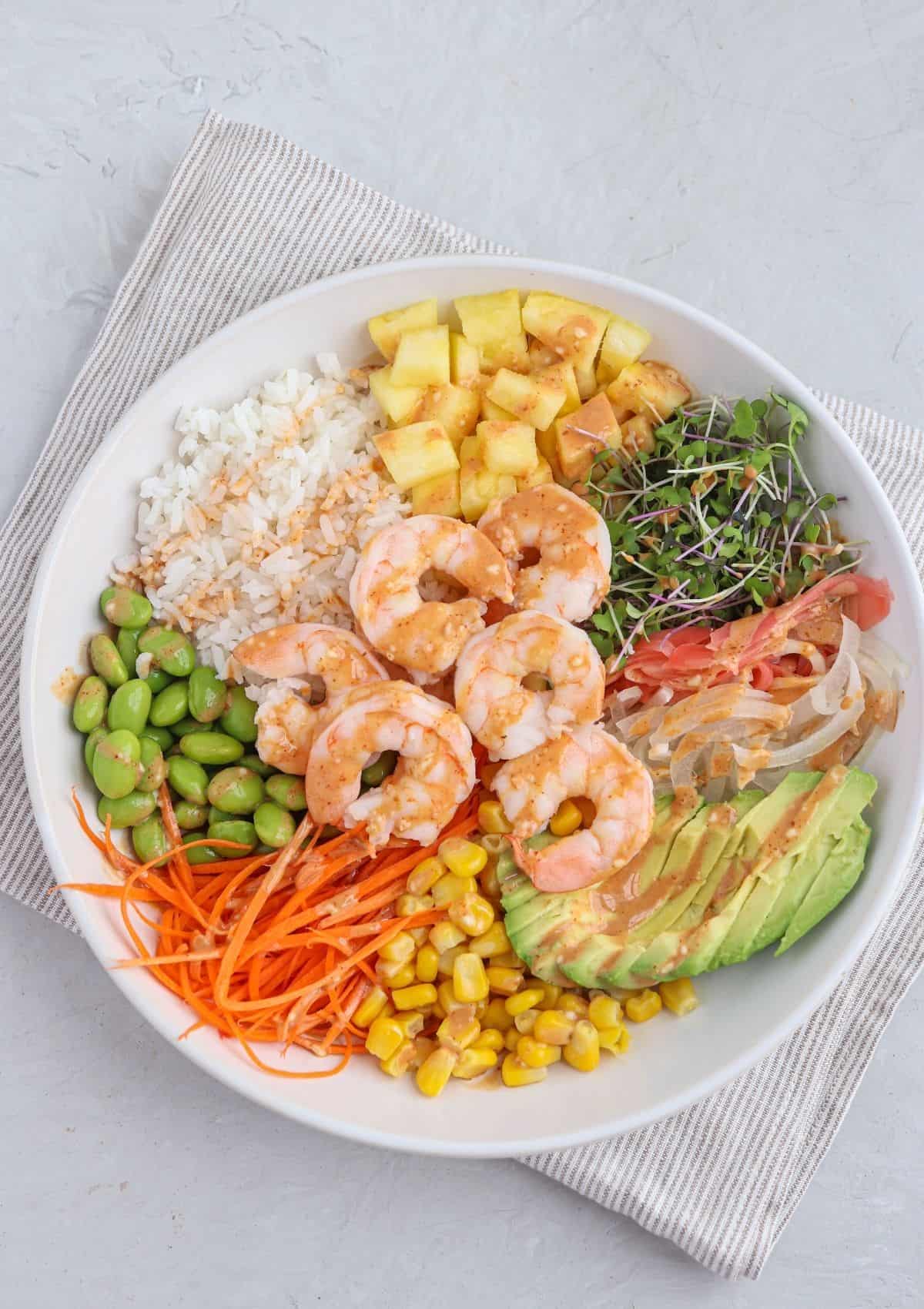 shrimp poke bowl in a white bowl with rice, pineapple, cooked shrimp, edamame, corn, carrots, microgreens and creamy spicy mayo.