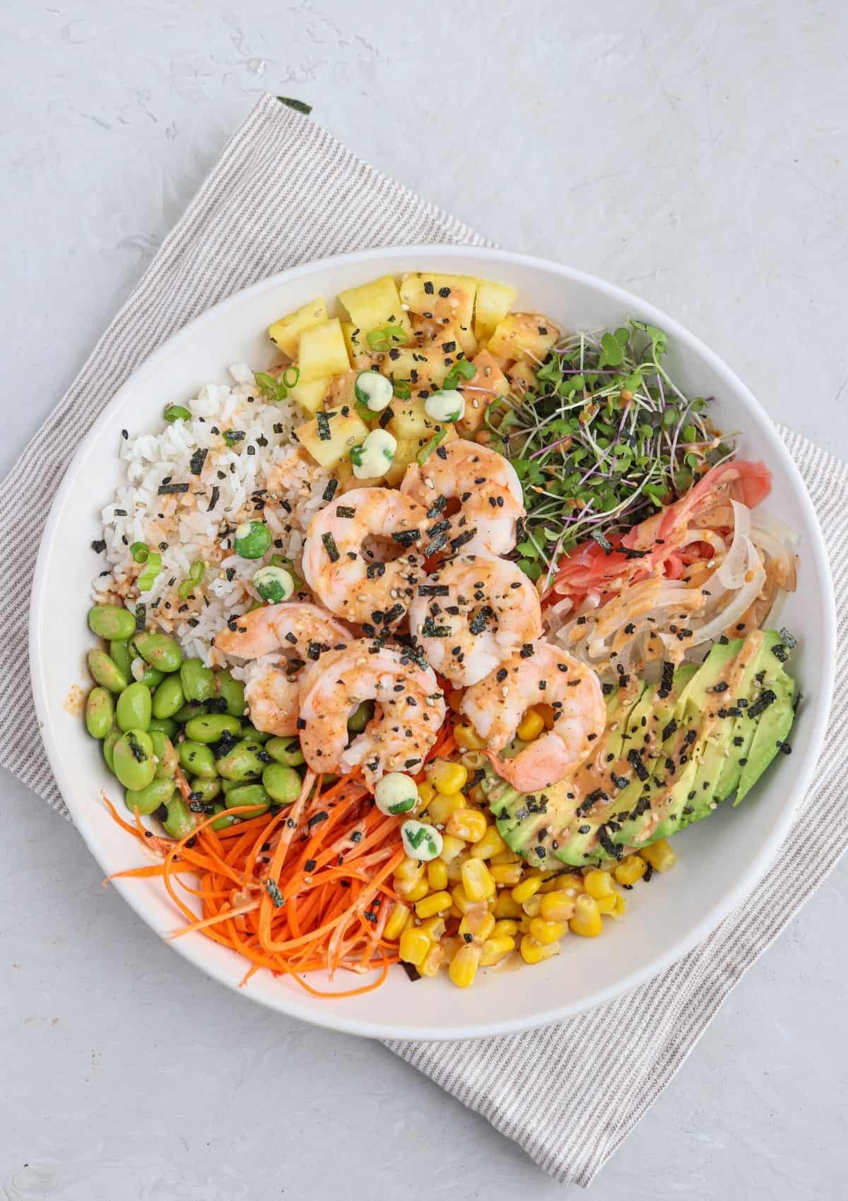 shrimp poke bowl in a white bowl with rice, pineapple, cooked shrimp, edamame, corn, carrots, microgreens and creamy spicy mayo.