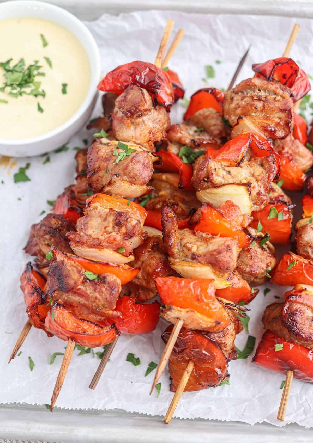 pork kebabs made with peppers, onions and bacon stacked on a baking sheet lined with paper with a side of mustard dipping sauce.