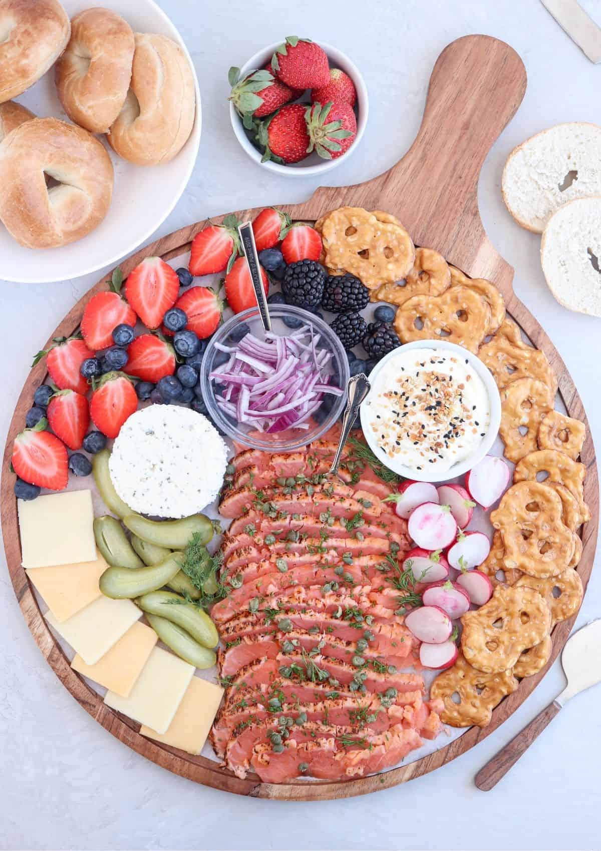 round wooden charcuterie board piled with smoked salmon, fruit, veggies, pickles, pretzel crisps and veggies.