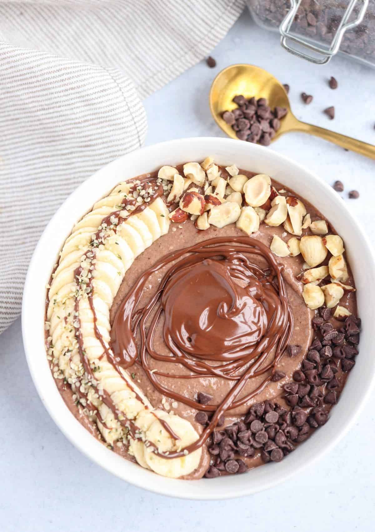 Nutella smoothie bowl in a white bowl topped with sliced banana, melted Nutella, toasted hazelnuts and mini chocolate chips.