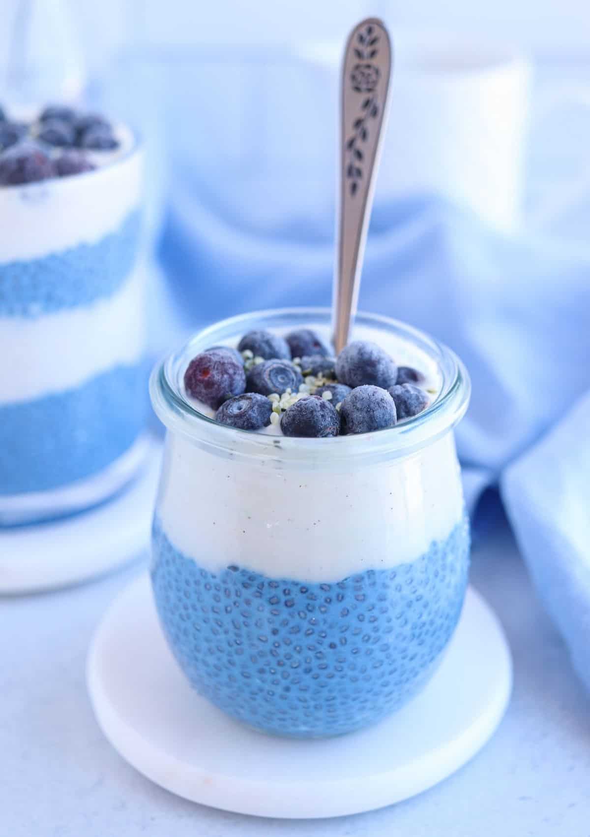blue chia pudding in a glass with a layer of yogurt and topped with blueberries and hemp hearts.