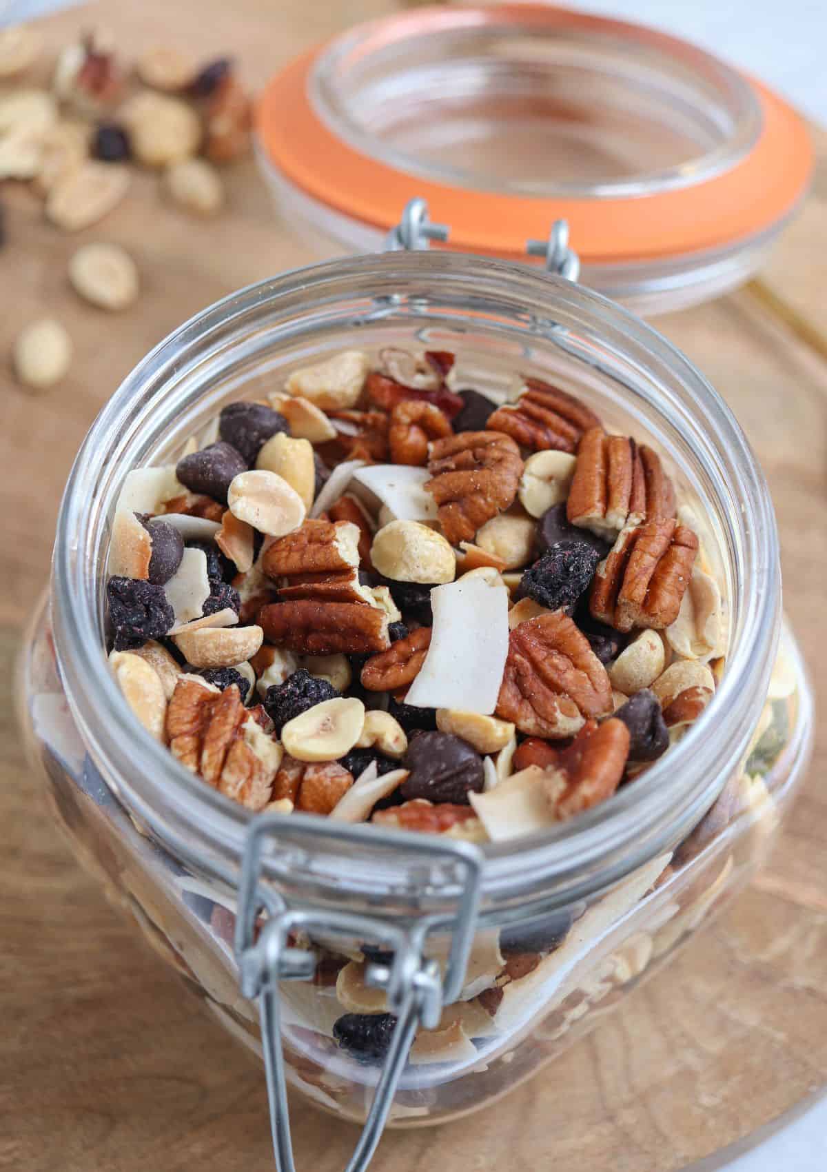 an open jar of low carb trail mix made with nuts, coconut chips, chocolate chips and dried blueberries on a wooden board.
