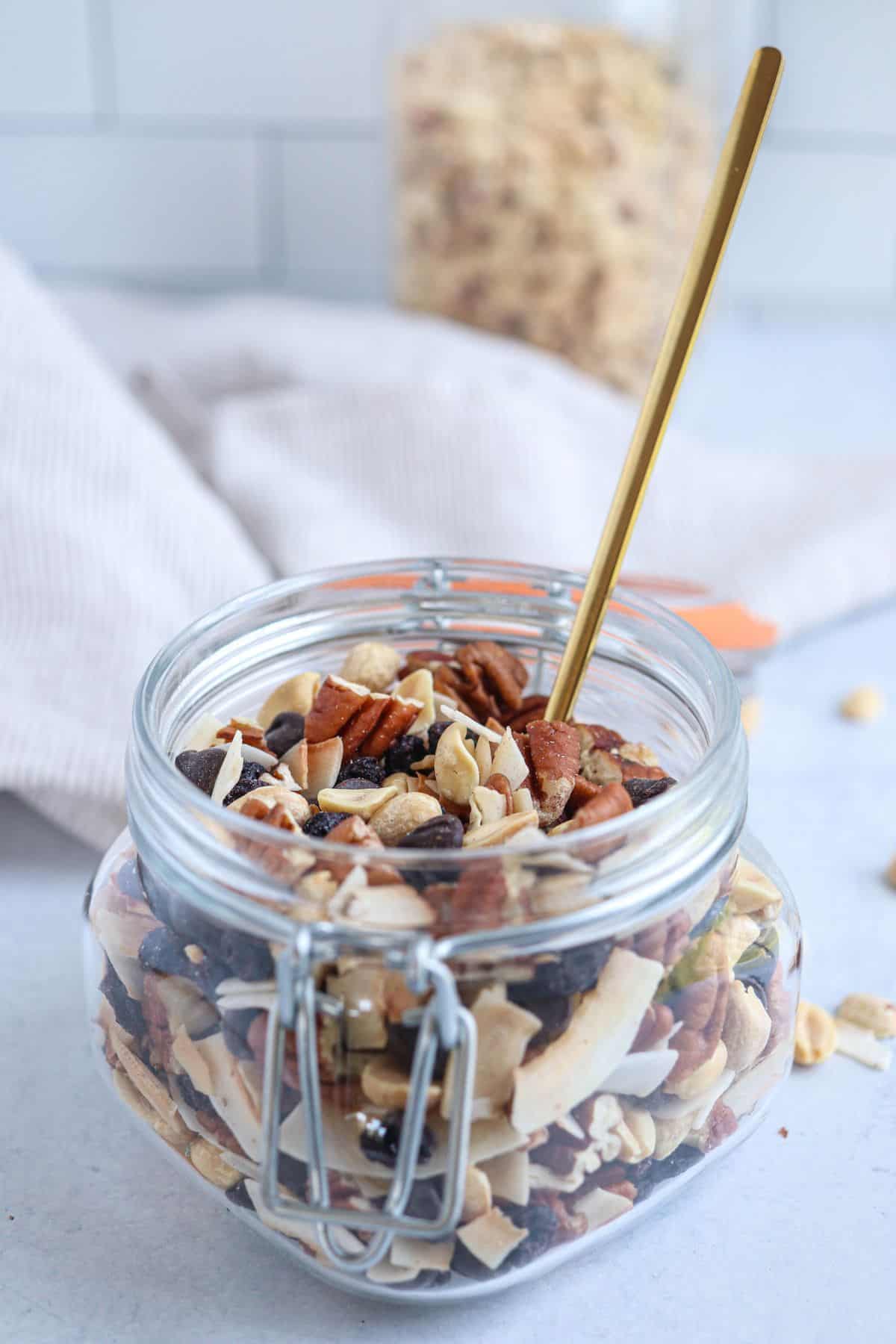 an open jar of low carb trail mix made with nuts, coconut chips, chocolate chips and dried blueberries on light gray counter with a golden spoon inside.