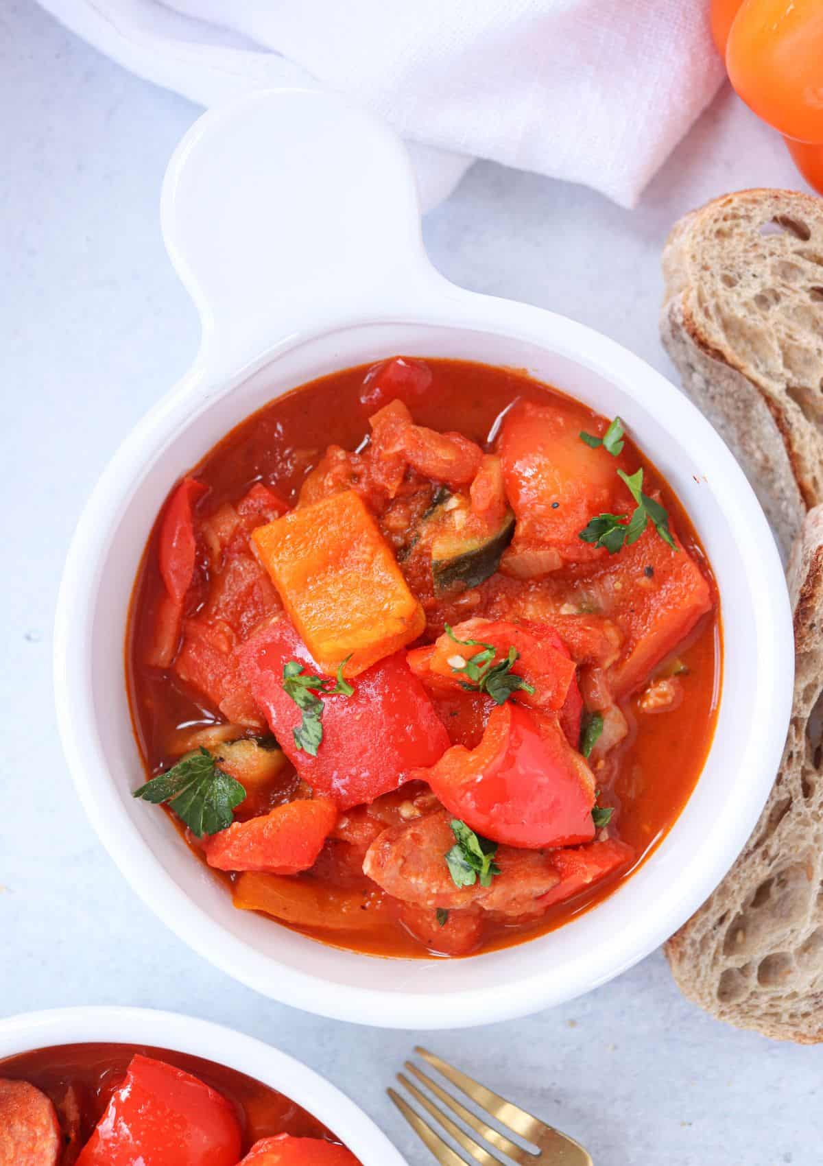 tomato pepper stew with sausage and zucchini in a white bowl with a side of crusty bread.
