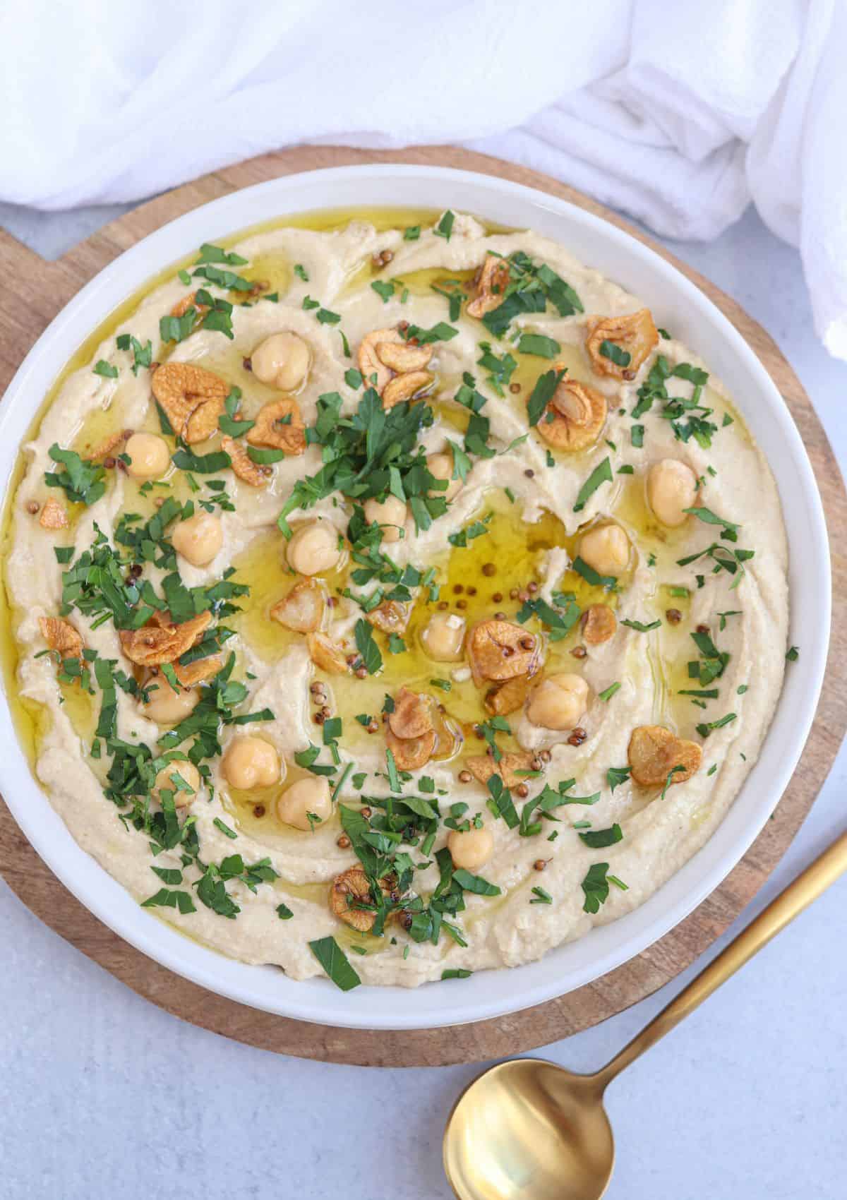 whole chickpea hummus in a white bowl topped with oil, chopped parsley, fried garlic, chickpeas.
