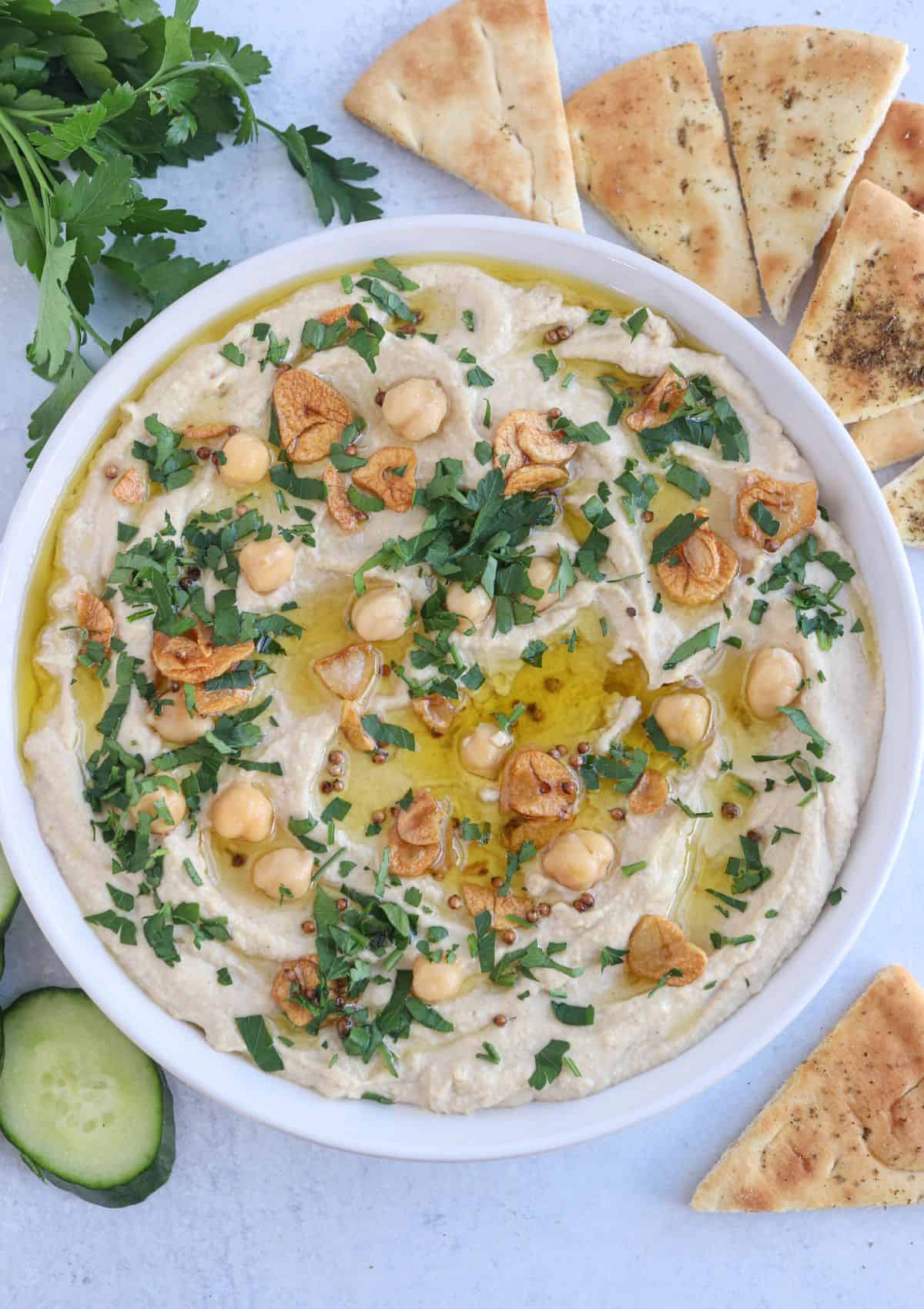 whole chickpea hummus in a white bowl topped with oil, chopped parsley, fried garlic, chickpeas with a side of pita chips, cucumber and parsley.