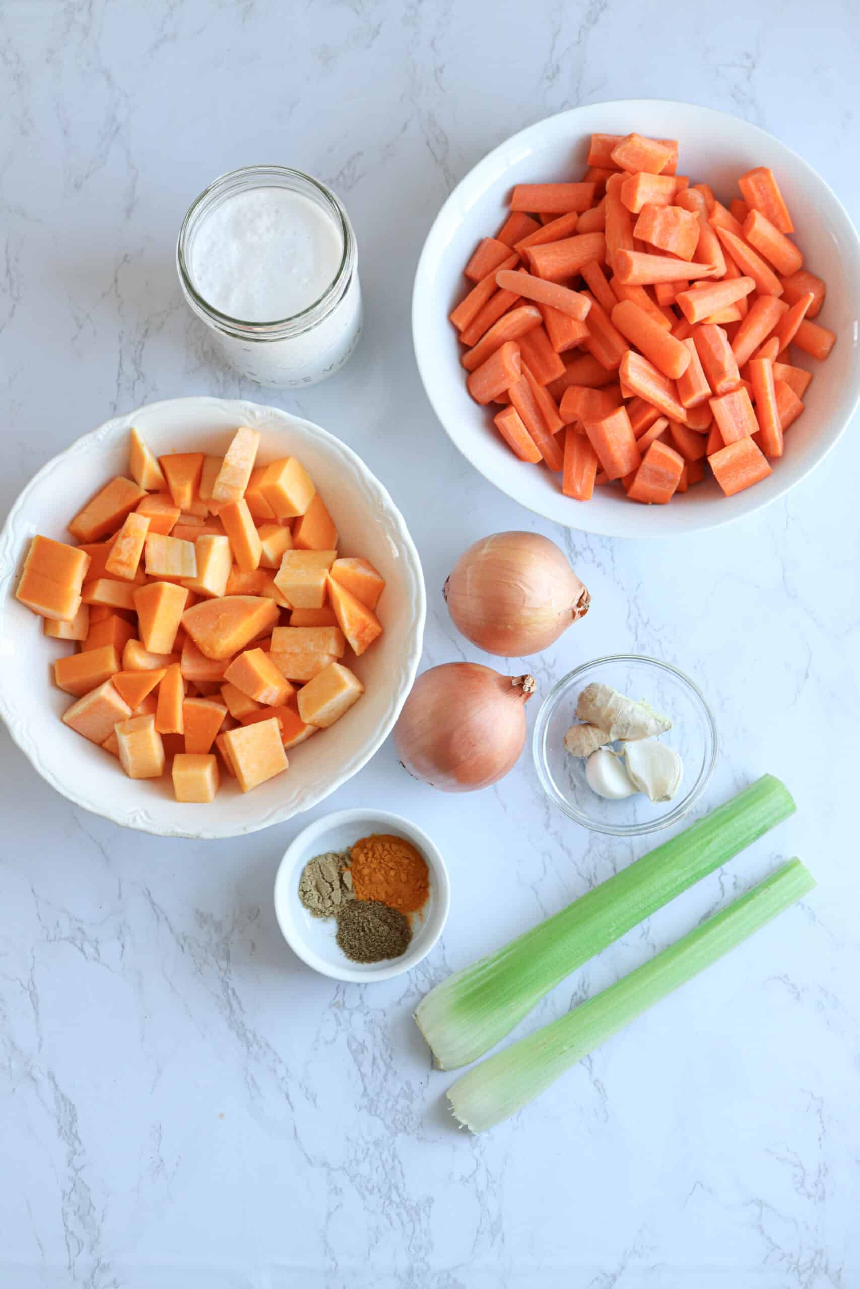 chopped butternut squash, carrots, two celery stalks, two onions, coconut milk, garlic and spices on white marble surface.