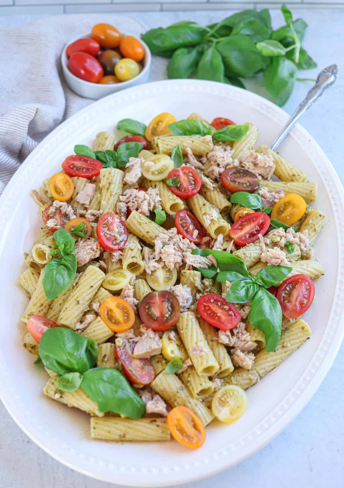 pesto tuna pasta with cherry tomatoes, tuna and fresh basil leaves on an oval white platter.