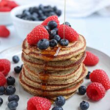 A stack of oatmeal protein pancakes topped with fresh raspberries and blueberries, with syrup being poured over them. More berries are scattered around the plate, and a bowl of blueberries is in the background.