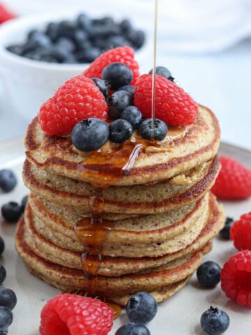 A stack of oatmeal protein pancakes topped with fresh raspberries and blueberries, with syrup being poured over them. More berries are scattered around the plate, and a bowl of blueberries is in the background.