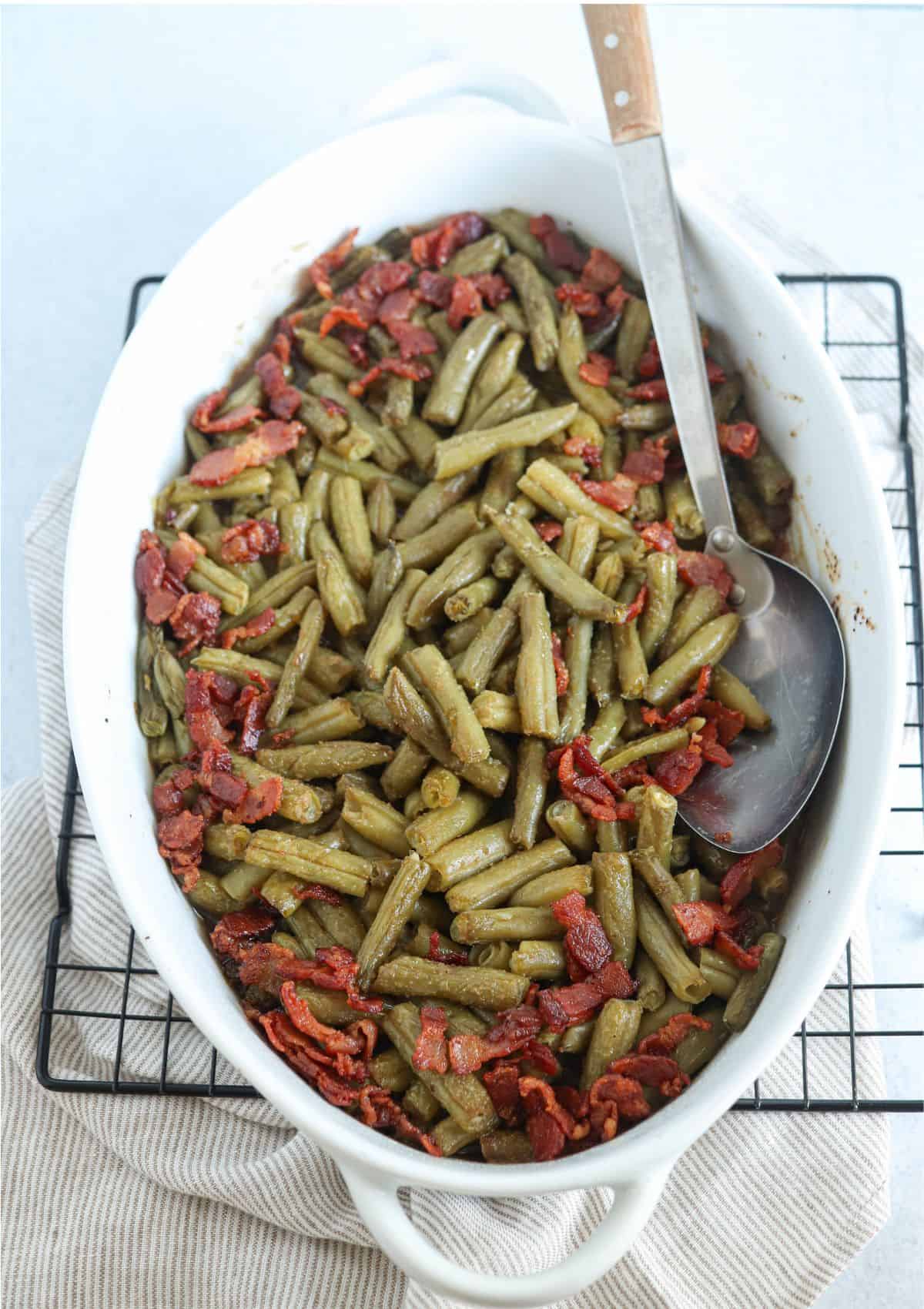 crack green beans in a white oval casserole dish topped with bacon bits  with a large silver spoon in on light gray surface.