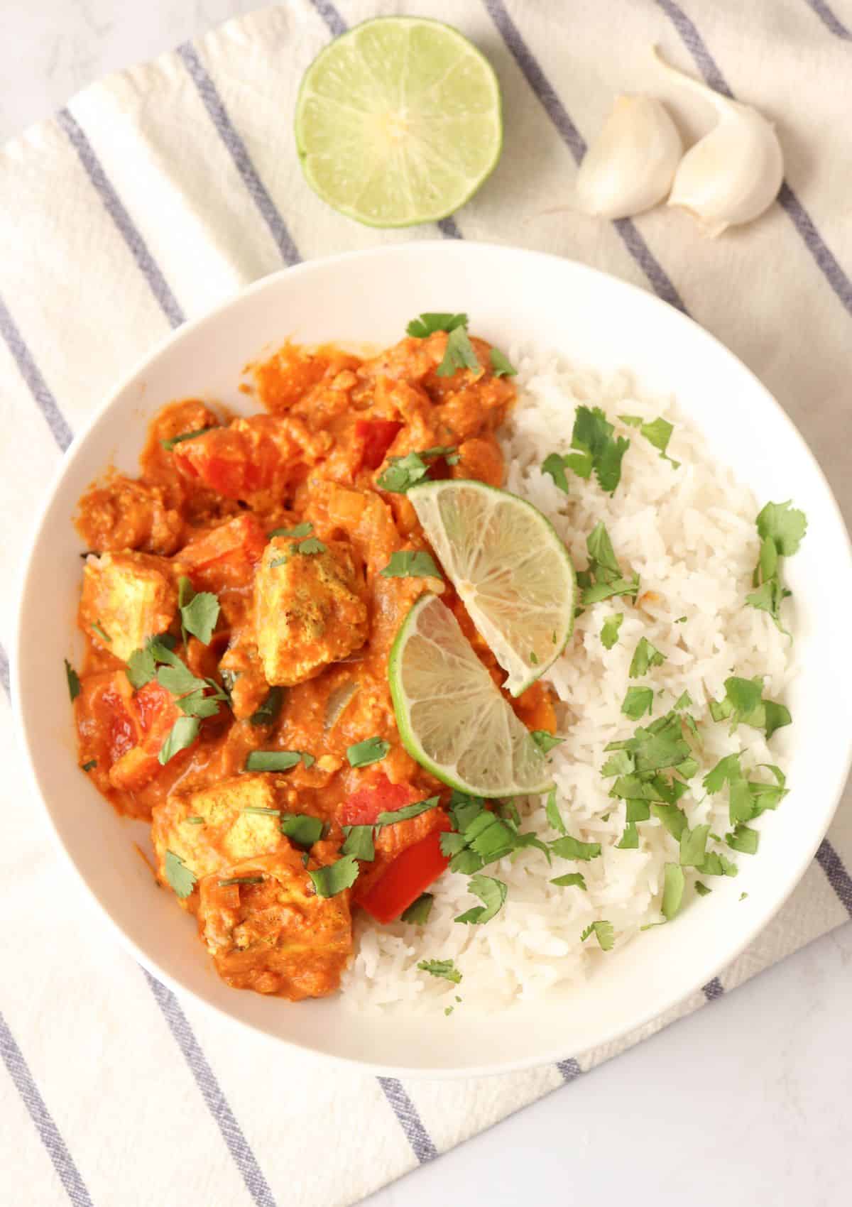 Indian tofu curry with a side of rice garnished with fresh cilantro leaves and lime slices in a white bowl.