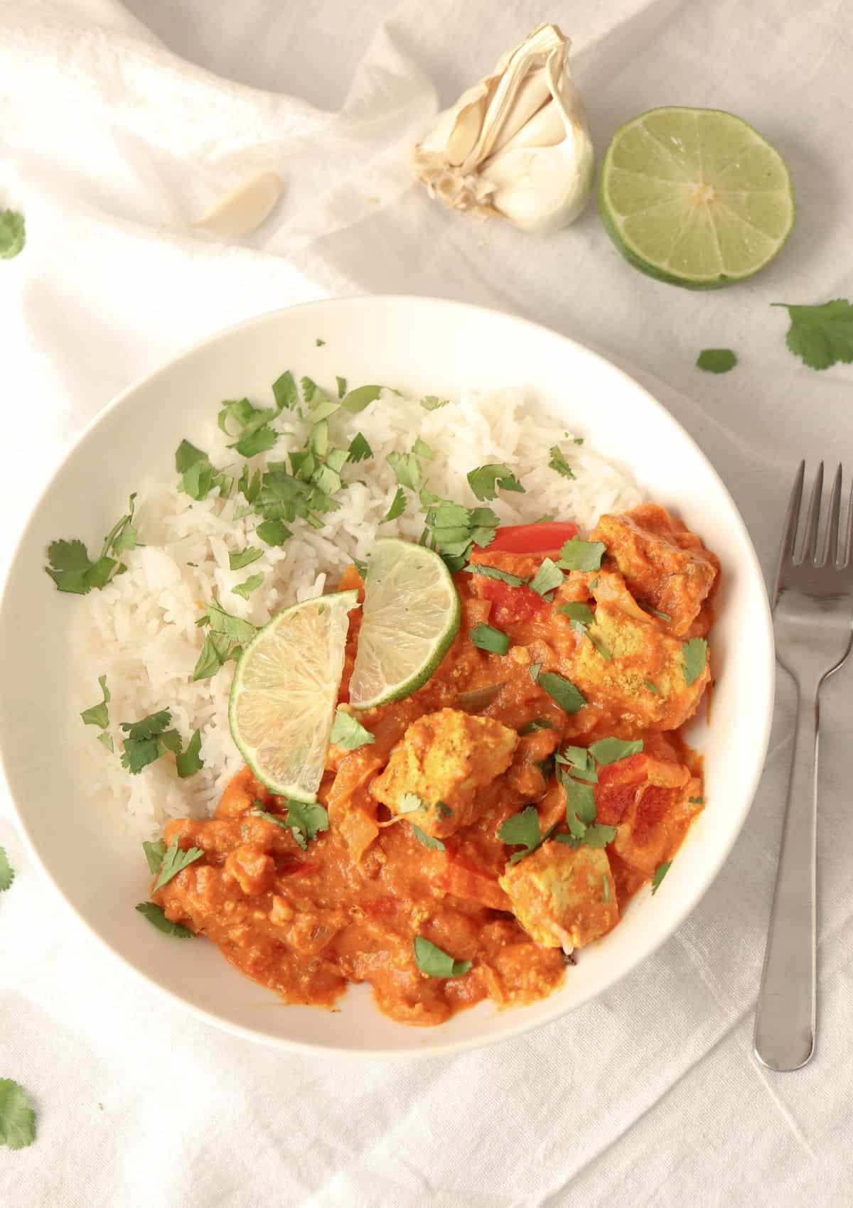 Indian tofu curry with a side of rice garnished with fresh cilantro leaves and lime slices in a white bowl.