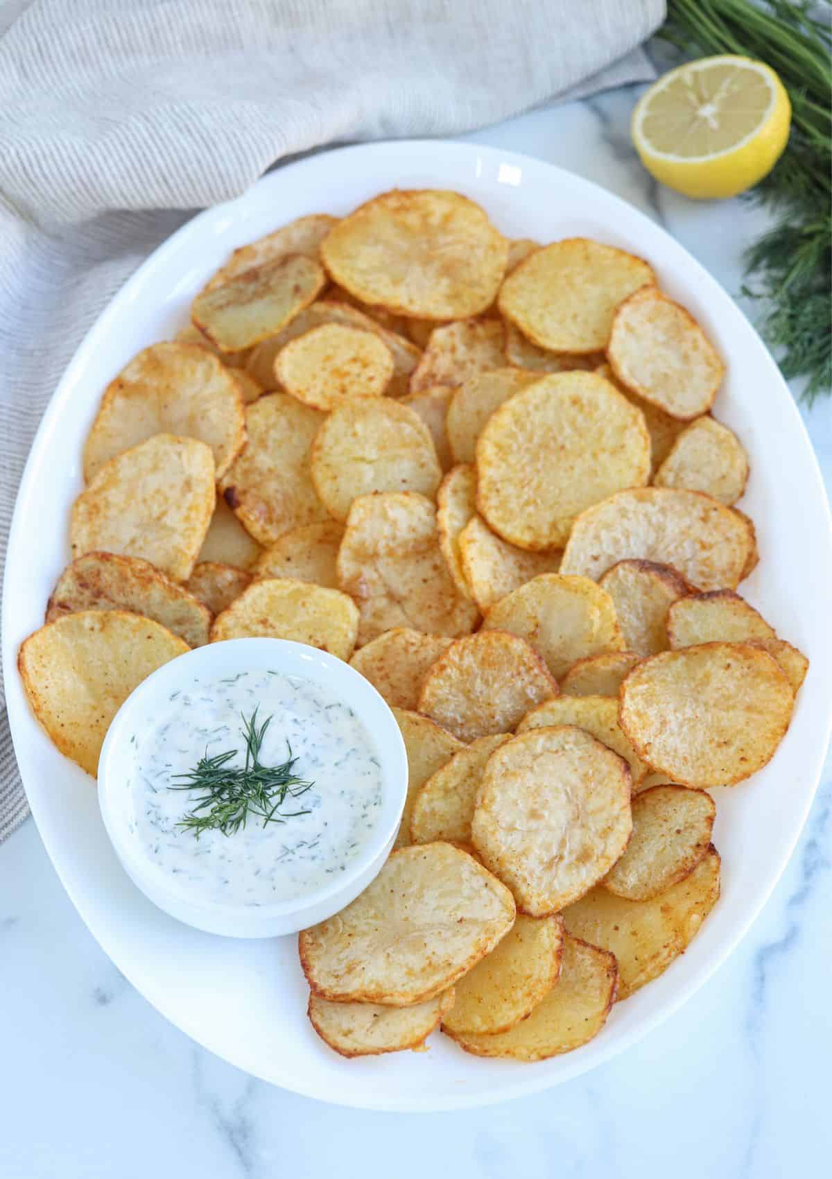 air fried potato slices on a white oval platter with a side of sill aioli in a small round white bowl. 