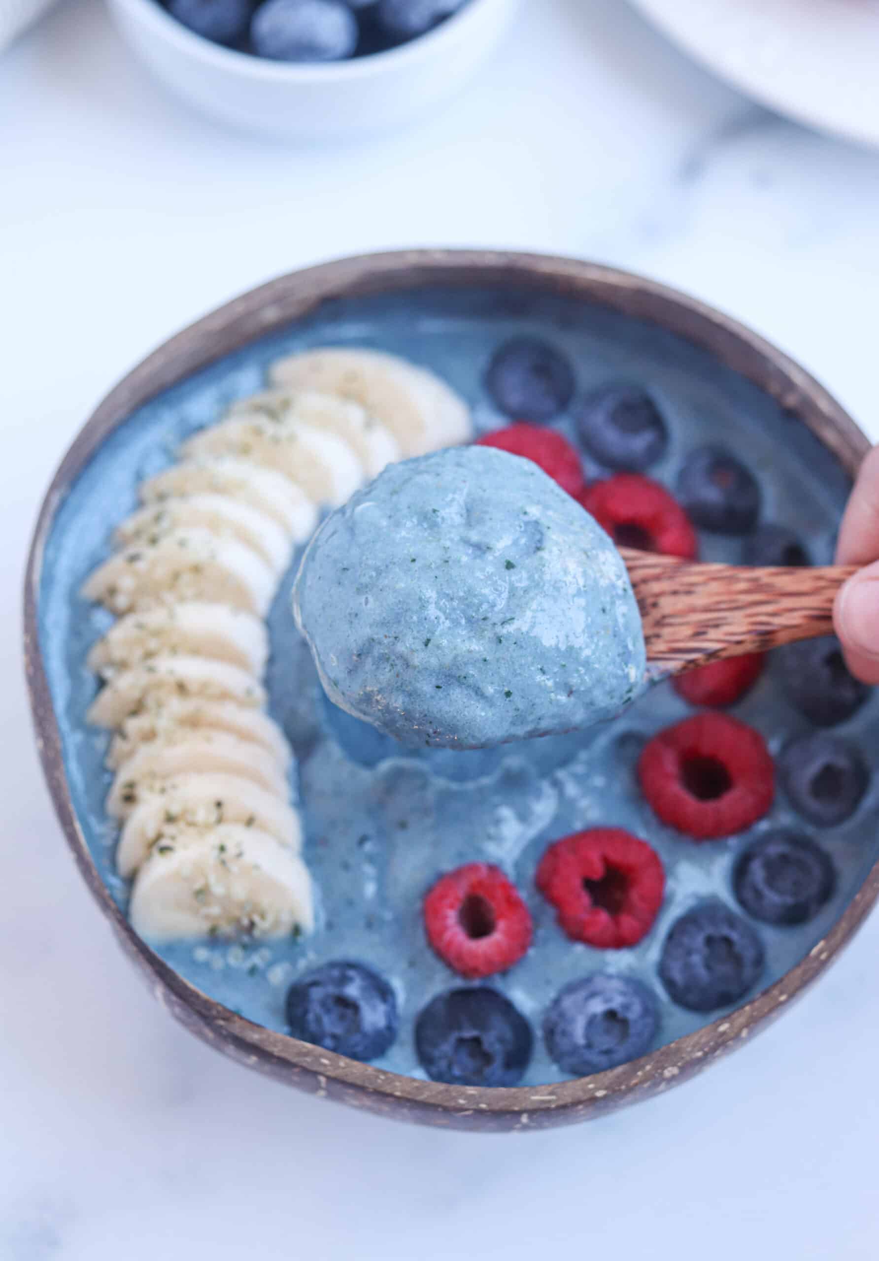 blue spirulina smoothie bowl in a coconut bowl topped with sliced banana, raspberries and blueberries, a spoonful view.