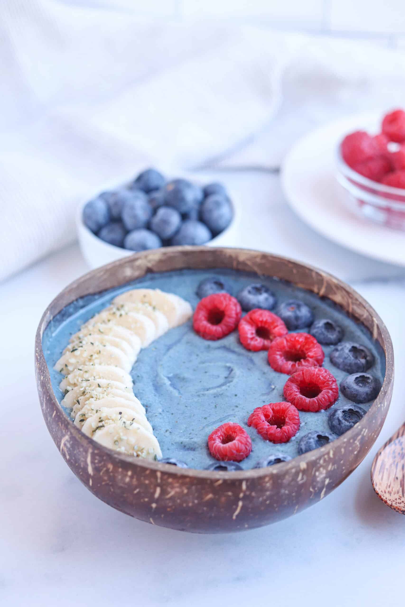 blue spirulina smoothie bowl in a coconut bowl topped with sliced banana, raspberries and blueberries.