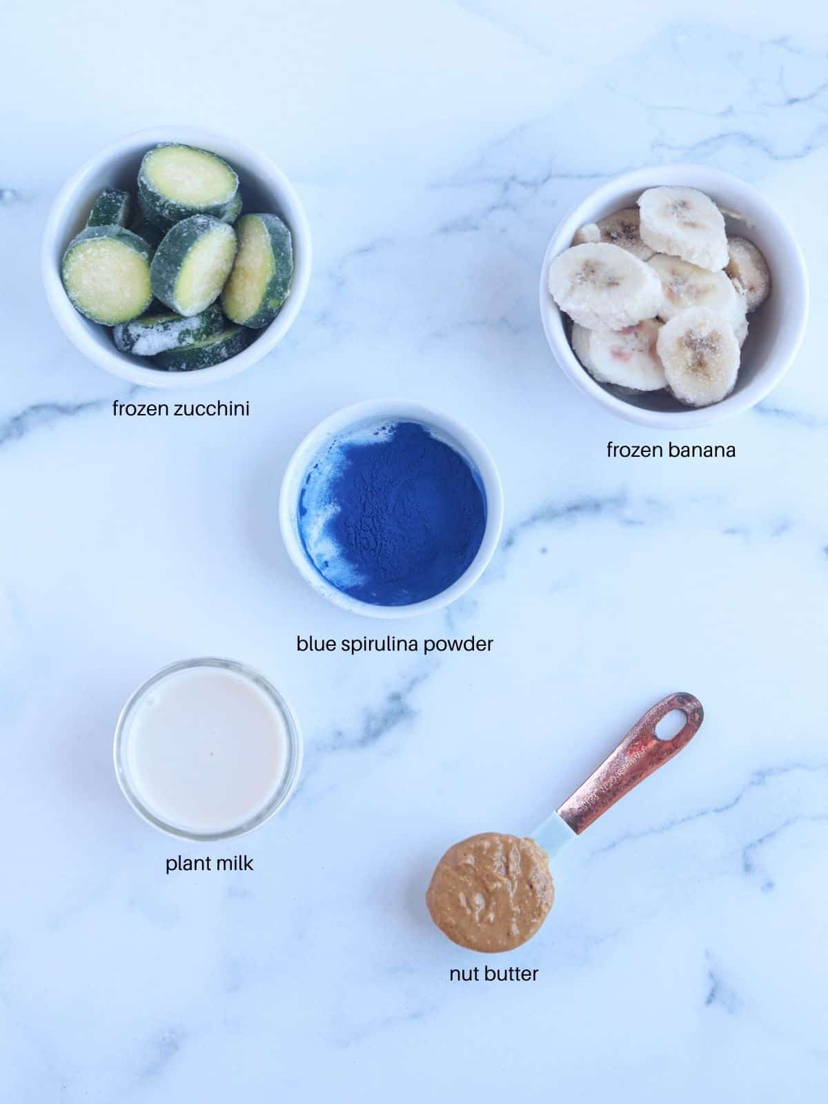 blue spirulina smoothie bowl ingredients in small round containers on white marble surface.