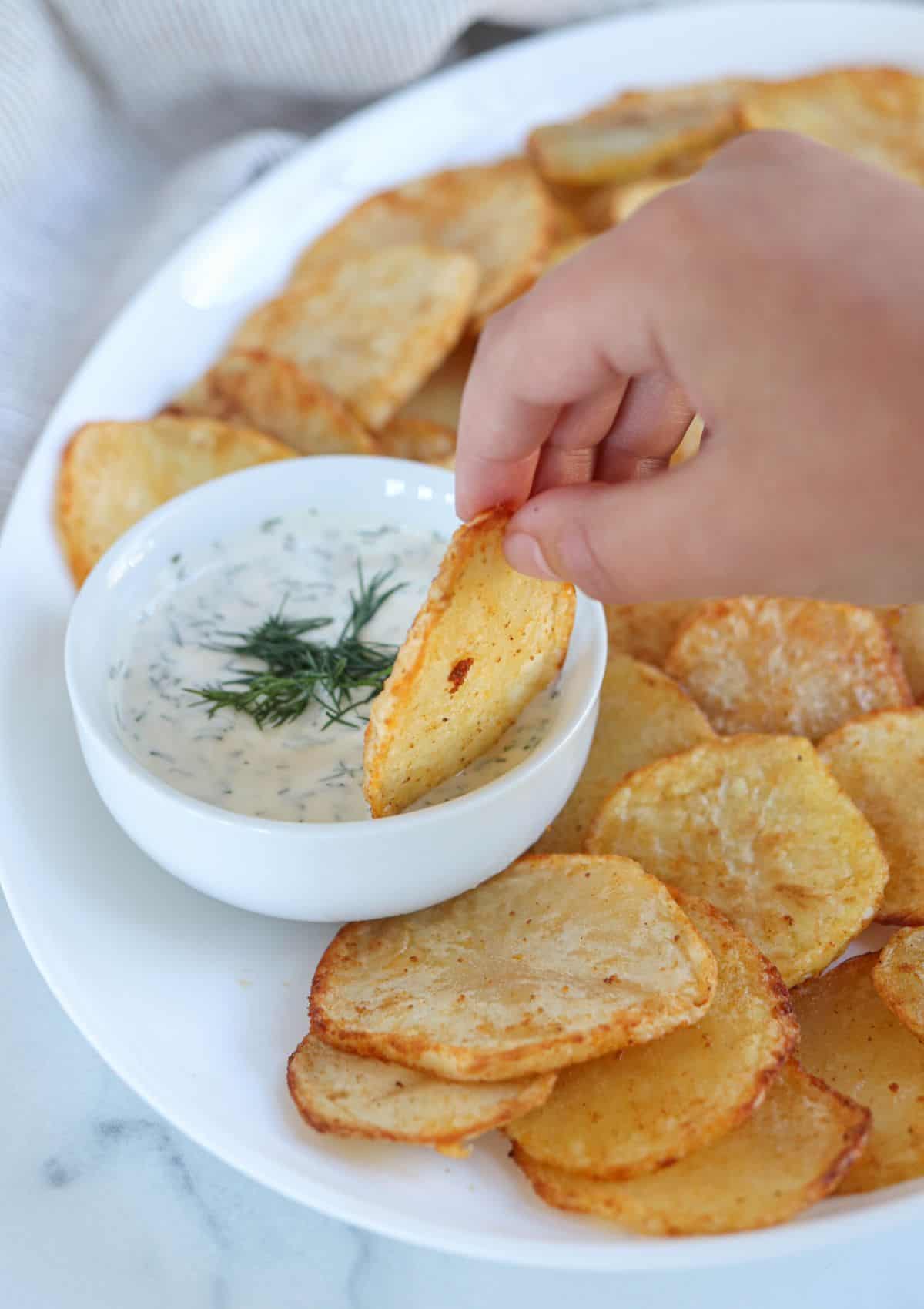 dipping air fried potato slice in dill aioli from a platter of crispy potato slices.