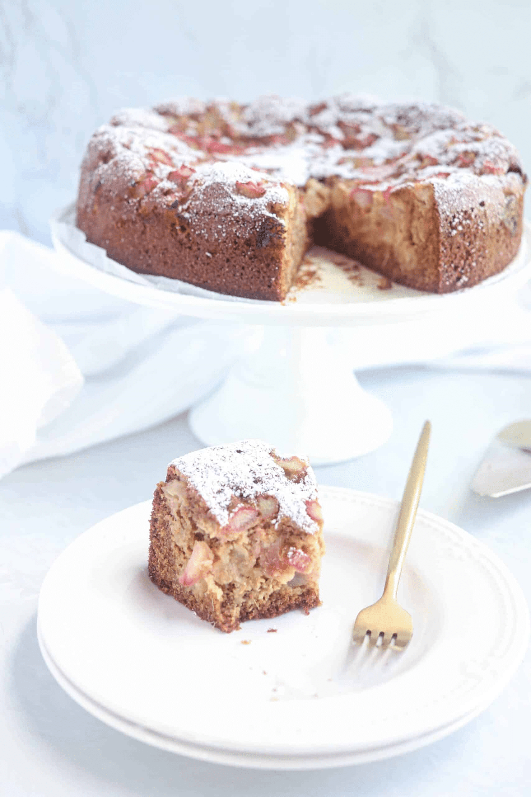 a piece of rhubarb cake on a white plate with the whole cake on a stand in the background, bite shot.