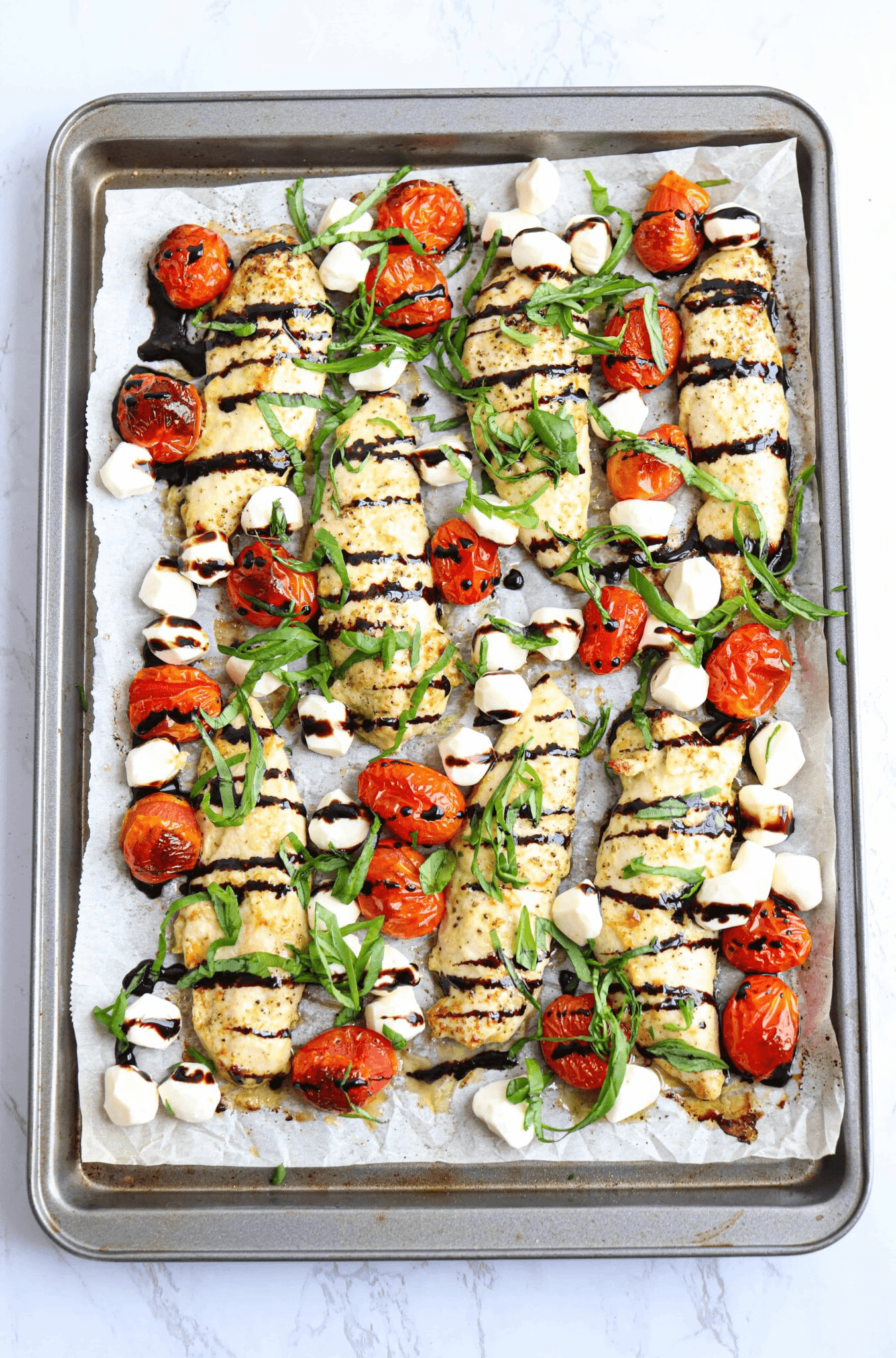 chicken tenders with mozzarella mini balls, roasted cherry tomatoes, fresh basil and balsamic drizzle on a paper lined baking sheet.