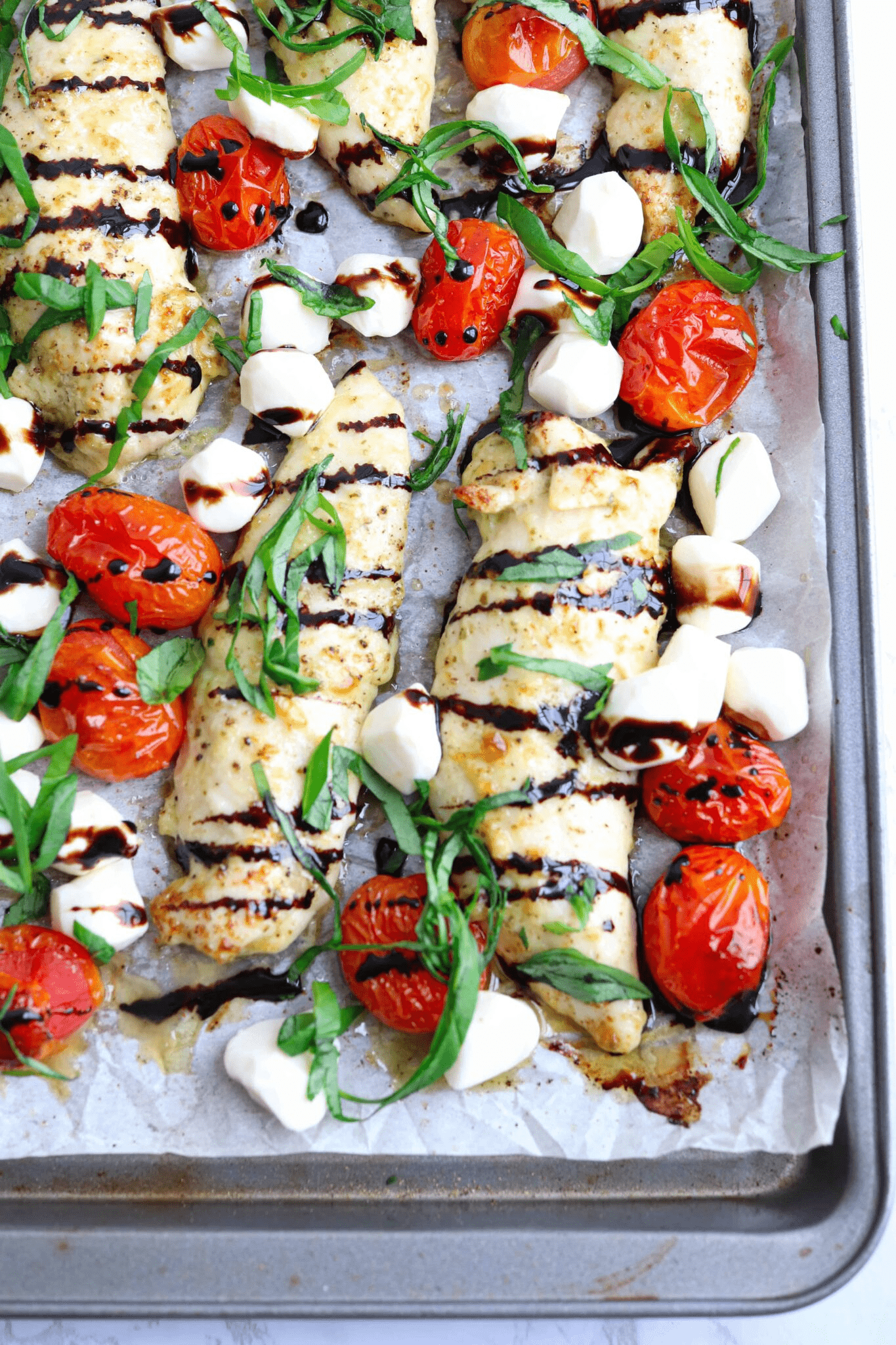 chicken tenders with mozzarella mini balls, roasted cherry tomatoes, fresh basil and balsamic drizzle on a paper lined baking sheet.