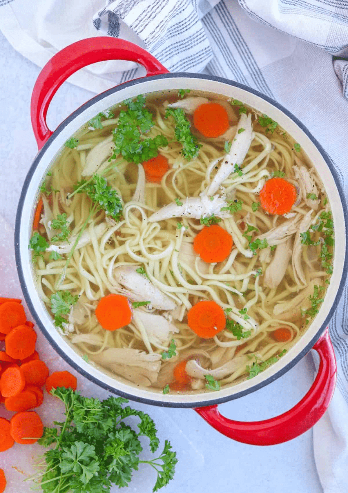 chicken noodle soup in a red Dutch oven garnished with carrot slices, chicken pieces and parsley.