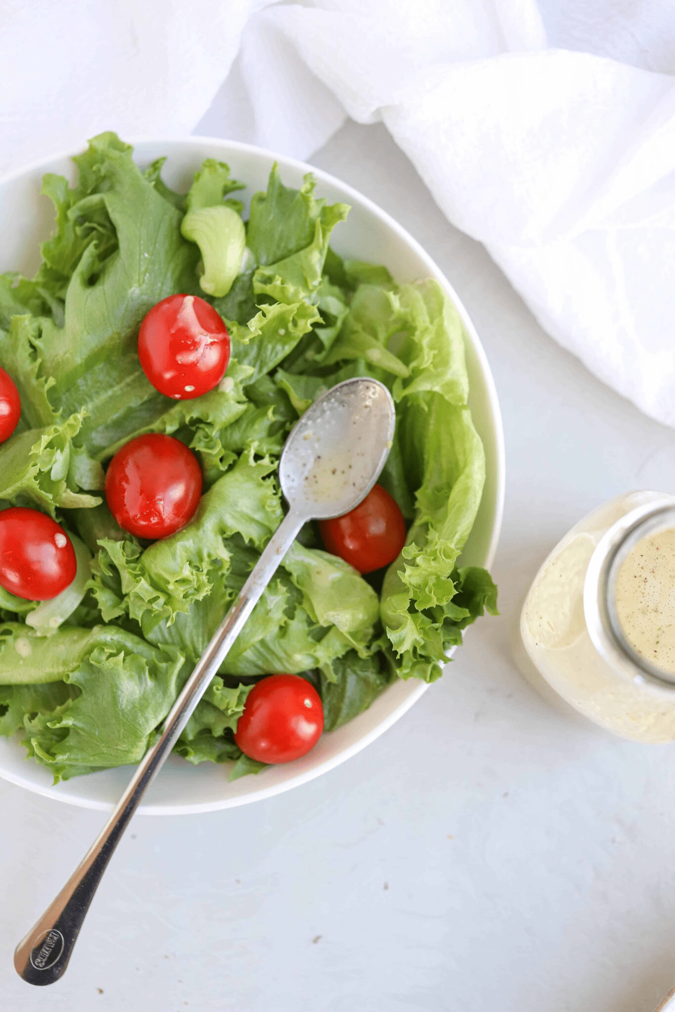 Green salad with cherry tomatoes in a white bowl with a long spoon and dressing next to it in a bottle.