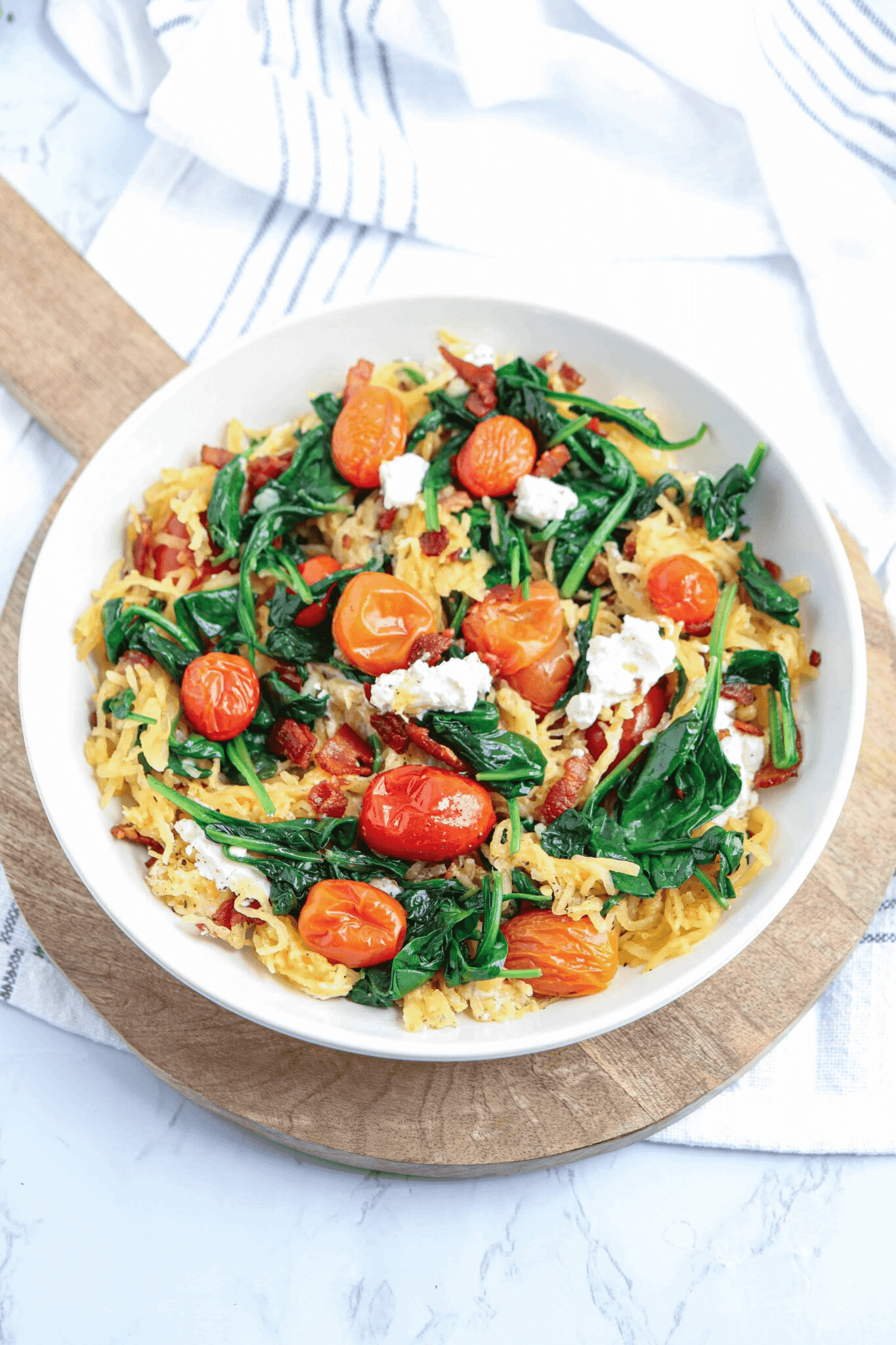 spaghetti squash with spinach, cherry tomatoes, goat cheese and bacon in a white bowl.